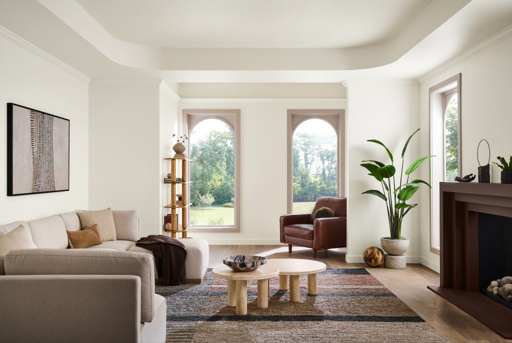 A warm, tonal living room with walls, ceiling, and baseboards painted Blank Canvas, window trim painted Mesa Taupe, and the fireplace mantel painted Baronial Brown. Layered warm neutrals, arched windows, soft upholstered seating, wood accents, and natural light create a relaxed, refined atmosphere.