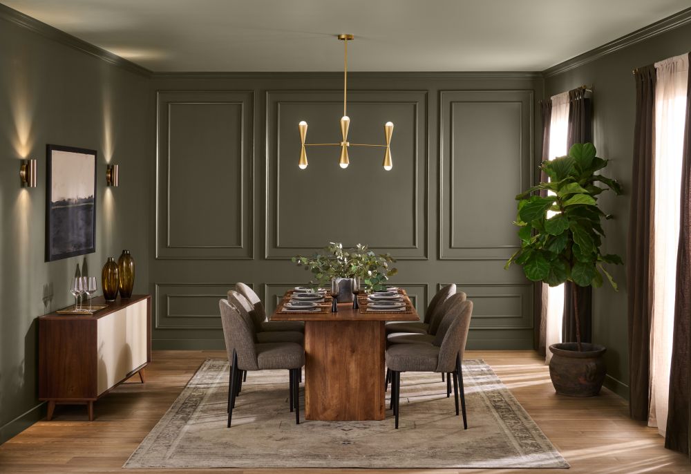 An elegant dining room with paneled walls painted BEHR Grounded, paired with an Environmental-painted ceiling. A warm wood dining table, upholstered chairs, brass chandelier, large potted plant, and tall windows create a sophisticated, nature-inspired setting.