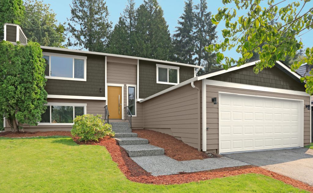 A suburban split-level Ranch style  home with shingles painted BEHR Grounded, siding in Introspective, and First Snow on the trim and garage door. A Radiance-painted front door adds a warm pop of color, while the manicured lawn, gravel walkway, and surrounding trees create a welcoming, nature-connected exterior.
