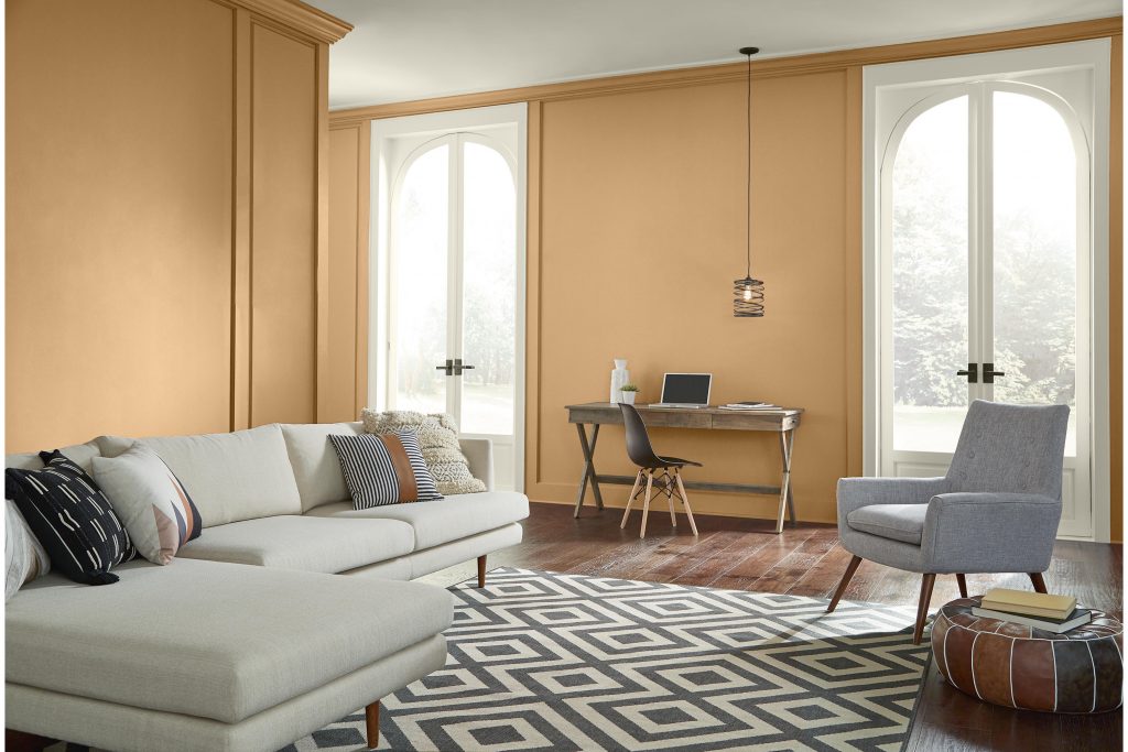 A bright living room with walls painted Cellini Gold and wood millwork painted Swiss Coffee, featuring tall arched windows, light neutral seating, a gray accent chair, wood flooring, and a geometric black-and-white rug.