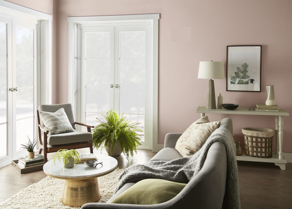 A bright living room painted in BEHR® Smokey Pink with crisp Ultra Pure White trim. Two large windows fill the space with natural light, while neutral furnishings, warm wood tones, and fresh greenery create a soft, airy feel.