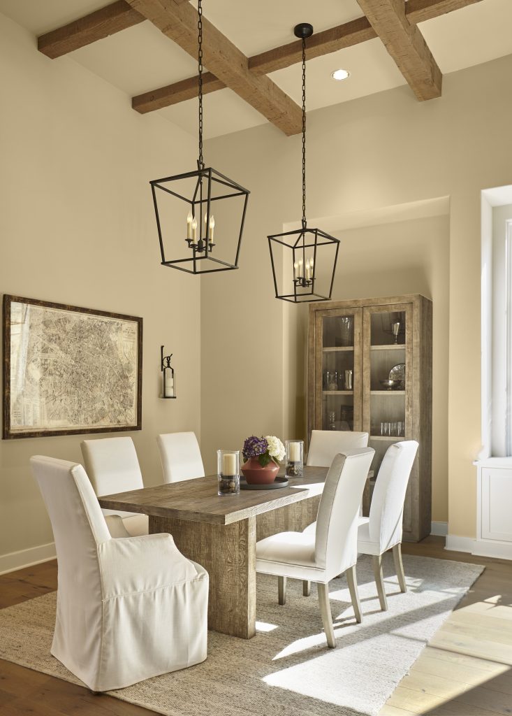 Dining room with walls painted Hayloft MQ2-22, a soft straw-gold paired with a rustic wood table, exposed beams, neutral seating, and black lighting.