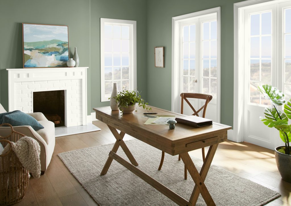 Sunlit home office with BEHR Bitter Sage N390-4 sage green walls, a white fireplace and a natural wood desk.
