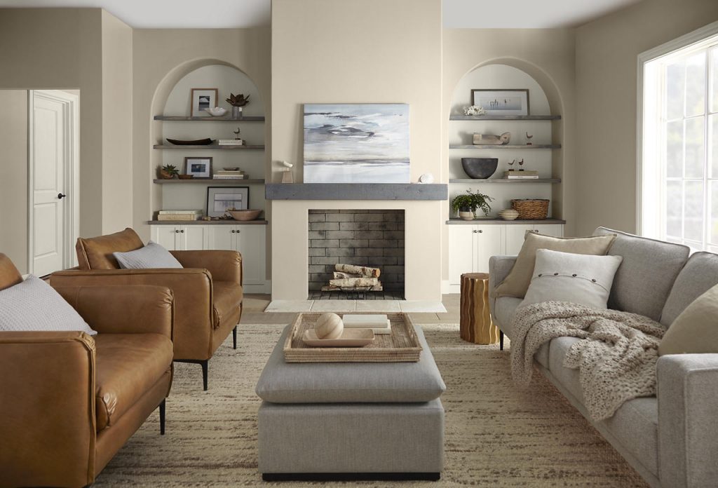 A warm, transitional living room with walls painted Even Better Beige and trim painted Blank Canvas. Camel leather armchairs, a soft gray sofa, arched built-in shelving, layered neutral textiles, and a simple fireplace create a comfortable, collected look. Abundant natural light and the warm beige-and-cream palette give the room an inviting, relaxed mood with subtle modern sophistication.