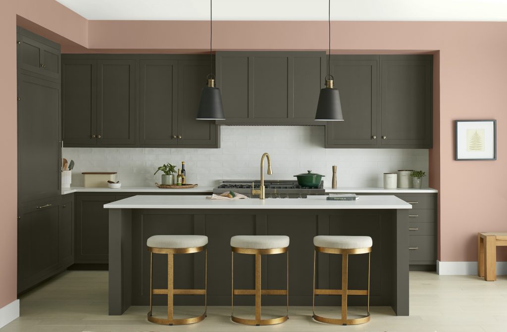 A contemporary kitchen with walls painted BEHR Sedona Pink, cabinetry and island in Grounded, and Whipped Cream on the ceiling and baseboards. Brass accents, black pendant lights, white countertops, and warm wood details create a polished, inviting space.