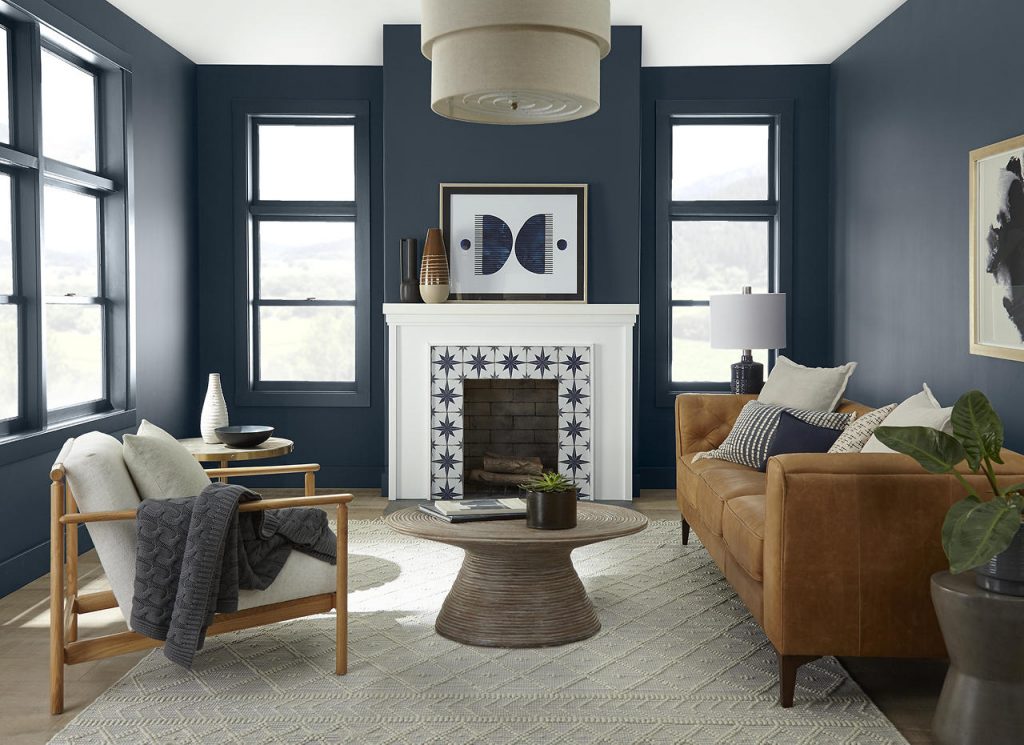 A casual living room with blue walls and trim painted Midnight Blue, a deep, moody navy blue, paired with ULTRA PURE WHITE® on the ceiling and fireplace. The space features a warm leather sofa, relaxed neutral seating, black-framed windows, natural wood accents, and a patterned tile fireplace surround.