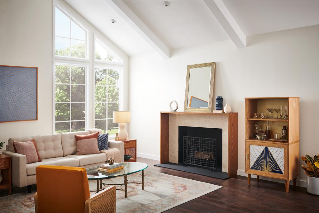 A bright mid-century modern living room with walls painted First Snow and trim and vaulted ceiling beams painted ULTRA PURE WHITE®. Large windows fill the space with natural light, while warm wood furnishings, a streamlined fireplace, neutral seating, and a burnt orange accent chair add inviting mid-century modern flair.