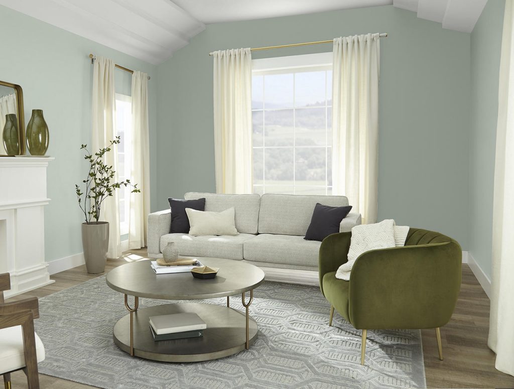 A bright living room with walls painted Riverdale and vaulted ceiling, beams, and trim painted Whipped Cream, featuring large windows, a light neutral sofa, an olive-green accent chair, white fireplace, and soft natural light.