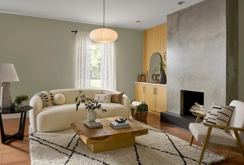 A contemporary living room with walls painted Jungle Camouflage and ceiling painted Silky White, featuring a modern plaster fireplace, warm wood built-ins, curved neutral seating, natural textures, and abundant daylight.