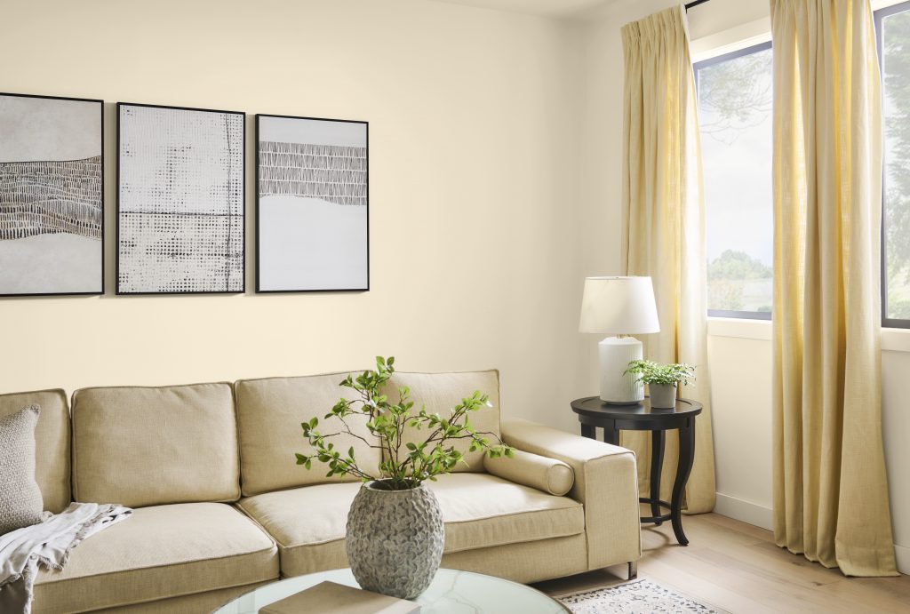 A bright living room with soft buttery yellow walls and trim painted Polished Pearl, featuring a beige sofa, large window with yellow tone curtains, neutral artwork, natural wood flooring, and fresh greenery.