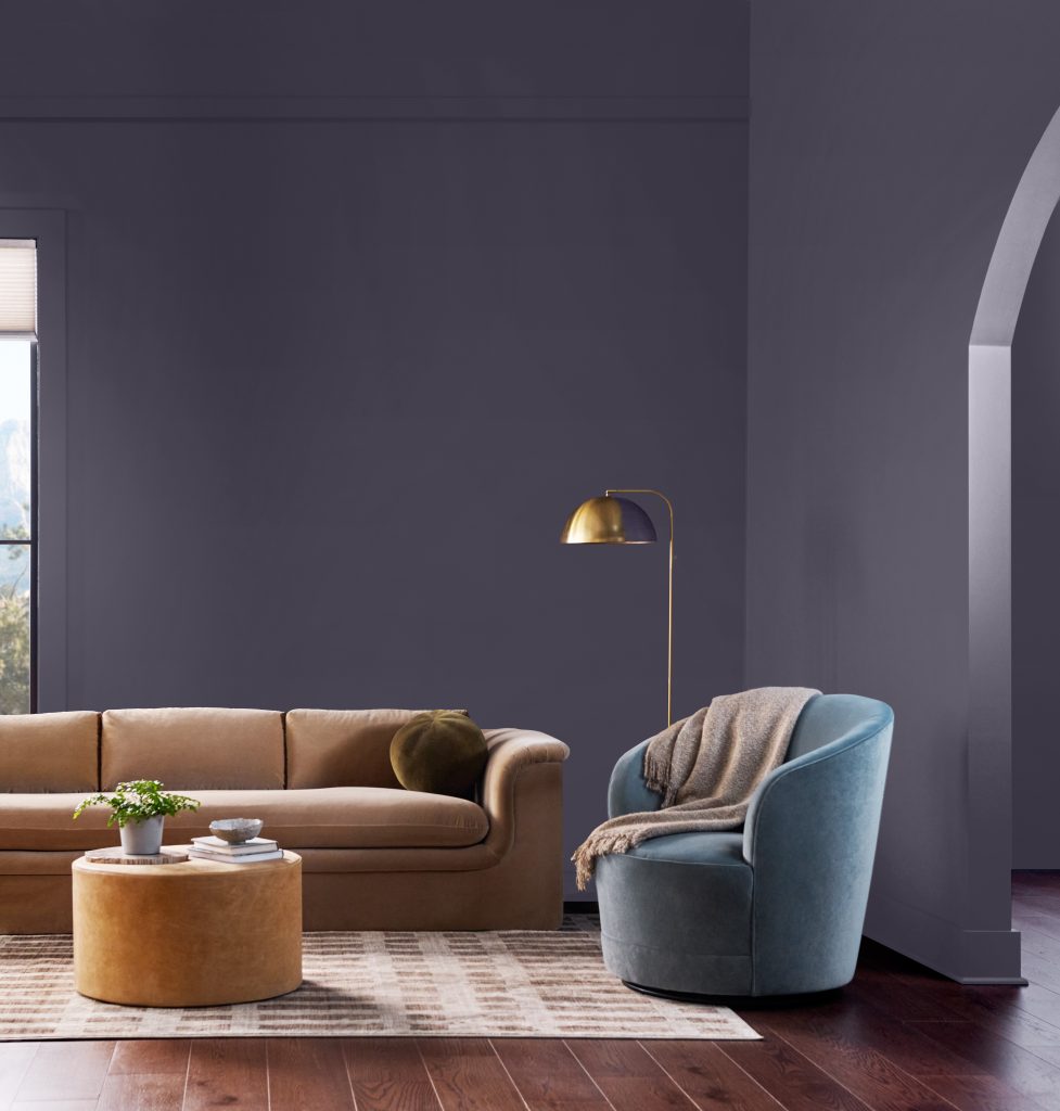 A contemporary living room with walls painted Plush Purple, a decadent, lush purple shade, featuring a warm camel sofa, soft blue accent chair, brass floor lamp, dark wood flooring, and a dramatic curved architectural opening.