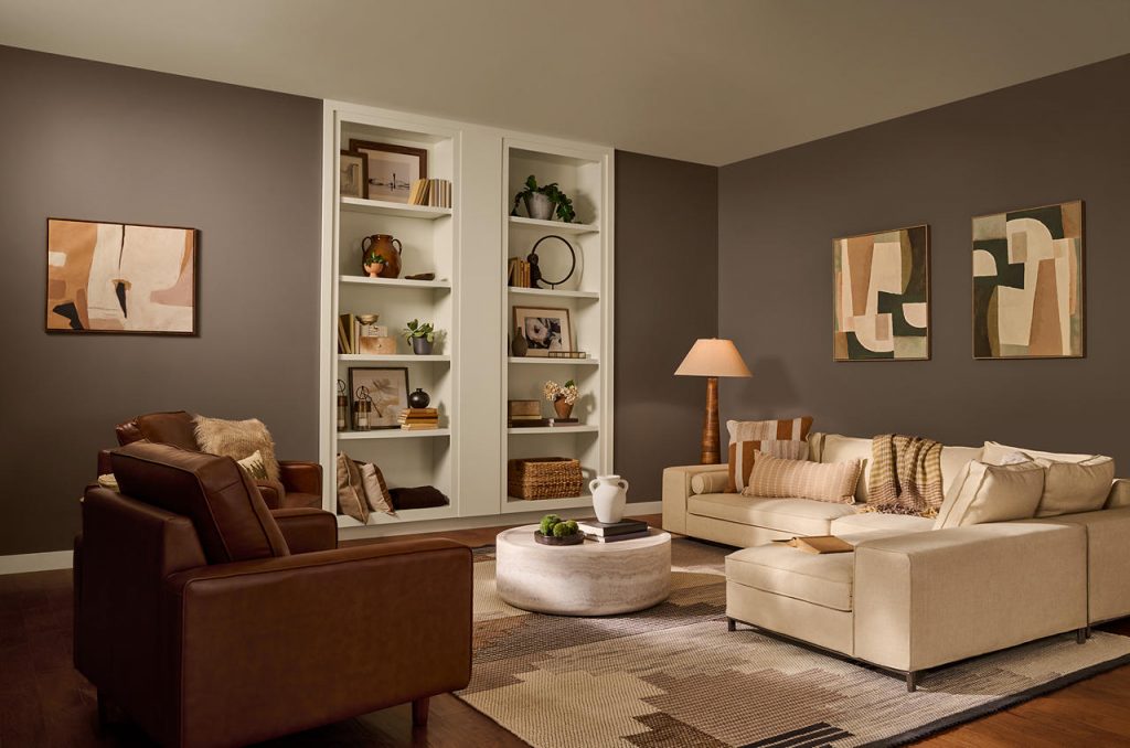 A warm, sophisticated basement-living room space with walls painted Introspective and the ceiling, trim, and built-in bookshelves painted Coastal Beige. Rich brown leather chairs, a soft cream sectional, layered neutral textiles, warm wood tones, and abstract artwork create a cozy, collected look. The tonal brown-and-beige palette, built-in shelving, and soft ambient lighting give the space a refined, intimate mood with a modern organic feel.