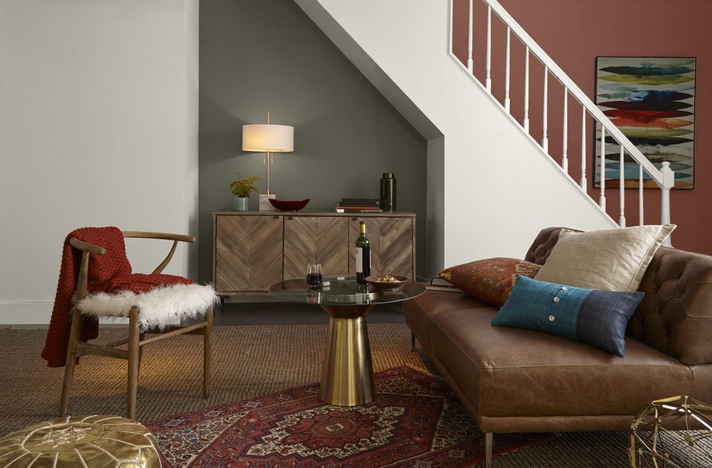 A cozy basement living area beneath a staircase with a Grounded-painted alcove, Painter’s White on the foreground wall and trim, and Tuscan Terra on the staircase back wall. A leather sofa, woven chair, patterned rug, wood cabinet, and warm metallic accents create a collected, inviting space.