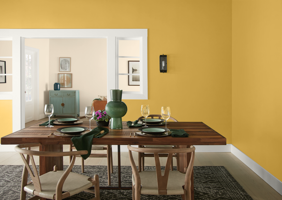 A contemporary dining room features a wooden table set with dark green dishes and glassware. The walls are painted a rich mustard yellow, contrasted by white trim and door frames. In the background, a sideboard and framed artwork decorate an adjoining room in softer neutral tones. Natural light and minimal decor create a clean, modern atmosphere.