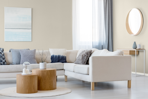A contemporary living room features a light beige sectional sofa accented with assorted throw pillows. Two round wooden coffee tables sit on a circular rug, holding minimalist decor pieces. In the background, a slim console table with a vase and a round wall mirror adds depth to the space. Soft curtains and a pastel abstract painting complete the airy, Scandinavian-inspired interior.