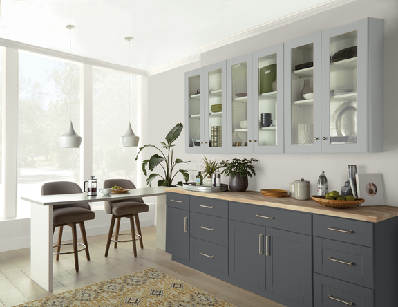 Contemporary kitchen interior featuring sleek gray lower cabinets and glass-front upper cupboards. A wooden countertop runs along the cabinetry, displaying plants, dishes, and small decor items. Large windows flood the space with natural light, illuminating a small dining area with two upholstered barstools and pendant lights. The overall palette is neutral with soft grays, whites, and warm wood tones, creating a clean and inviting atmosphere.