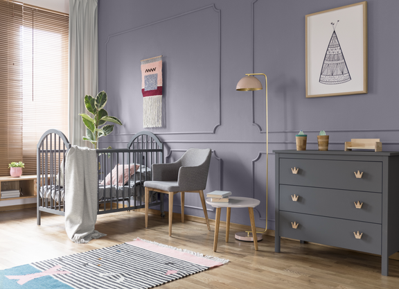 A modern nursery features a dark gray crib against a muted lavender wall with decorative molding. A cozy armchair, small side table, and floor lamp create a comfortable seating area beside the crib. On the right, a gray dresser with crown-shaped handles holds decorative items and pastel-colored cups. Soft textiles, a patterned rug, and natural wood flooring add warmth to the serene, contemporary space.