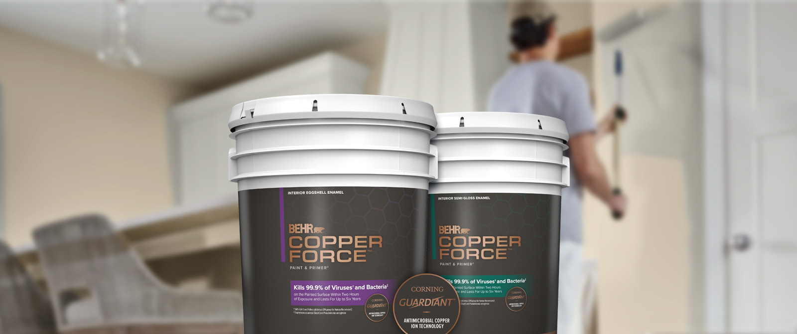 A paint contractor painting a wall in the background with two 5 gallon cans of BEHR® COPPER FORCE™ Interior Paint in the foreground.